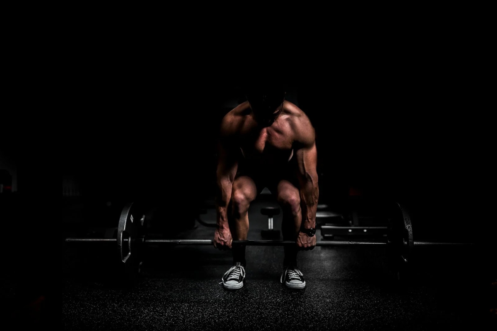 A muscular shirtless man grips a loaded barbell in a dark gym, ready to lift