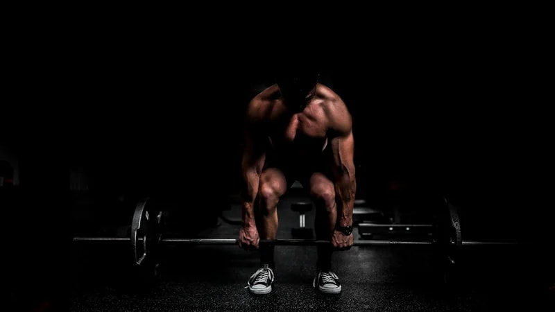 A muscular shirtless man grips a loaded barbell in a dark gym, ready to lift