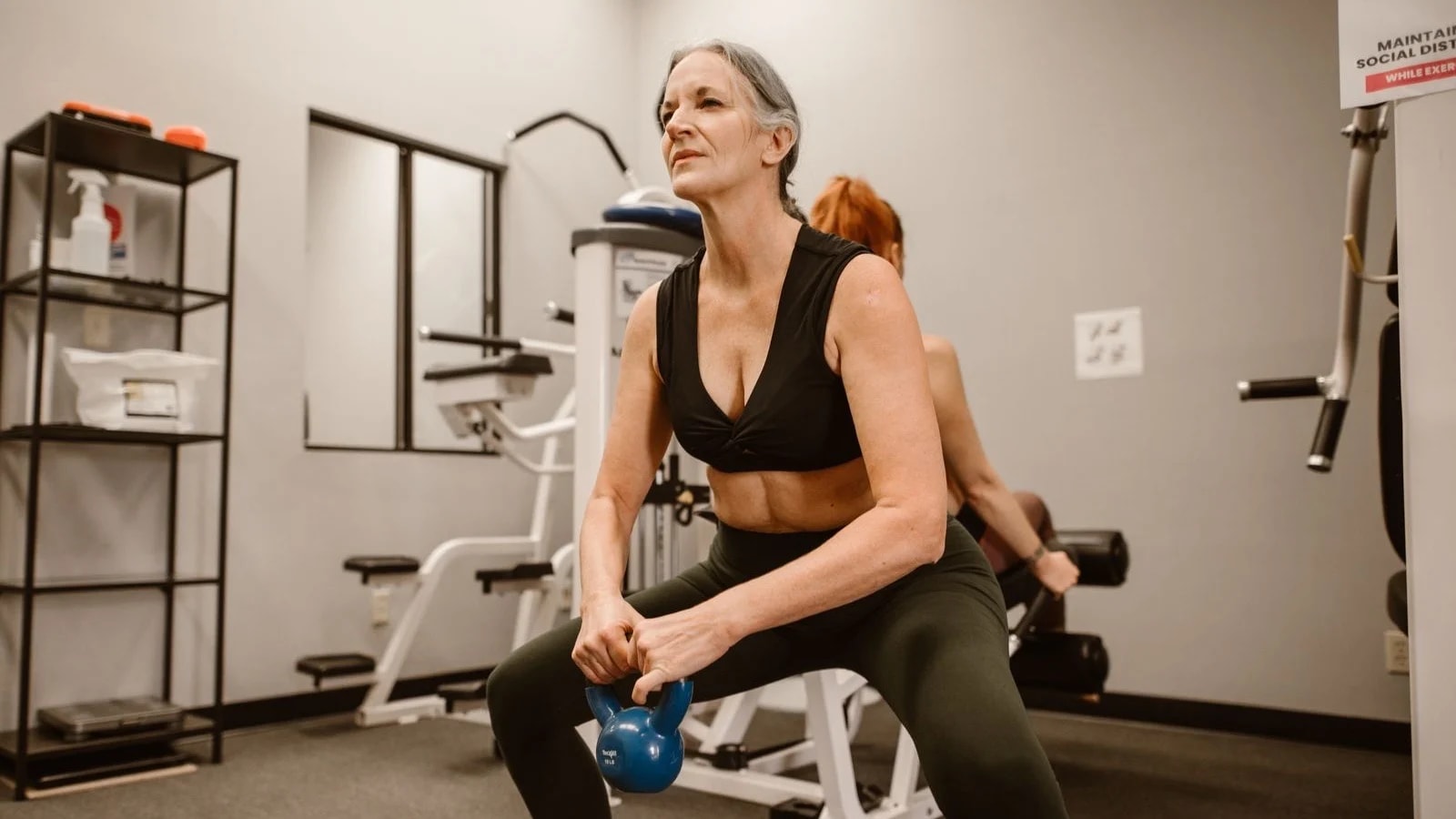 A woman with grey hair doing a kettlebell squat in a small gym