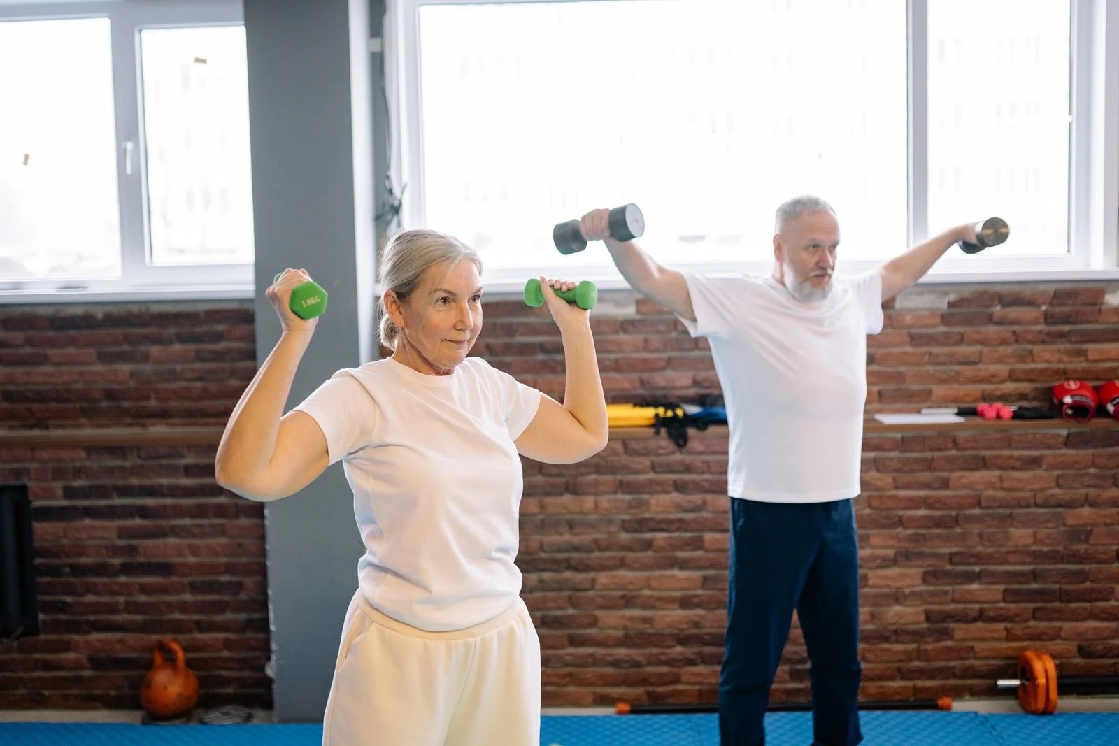 An older woman and man pressing light dumbbells overhead in a fitness studio