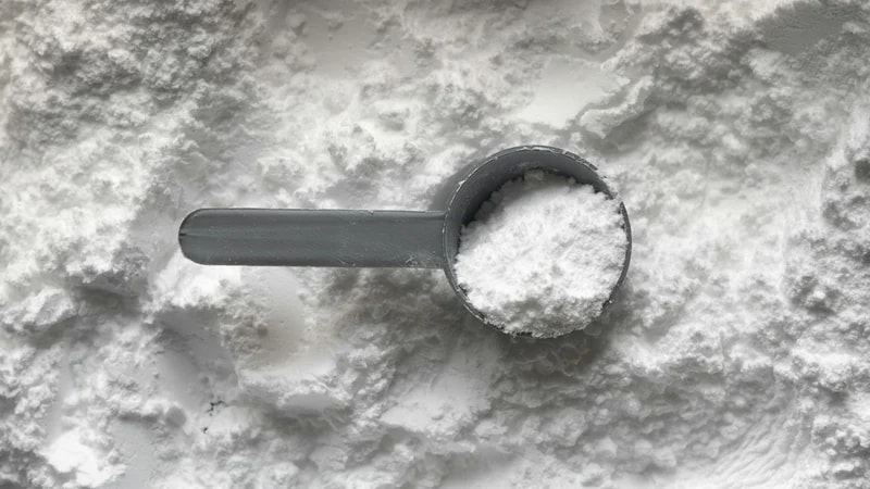 A grey plastic scoop filled with white powder resting on a bed of powder