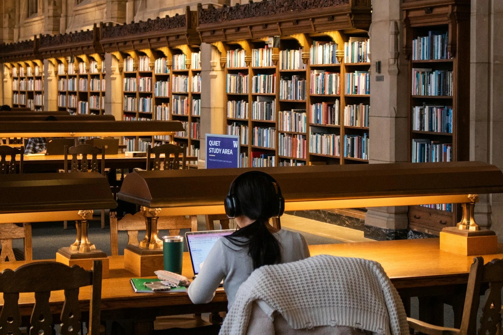 A student with headphones works on a laptop at a long wooden table in a quiet library study area