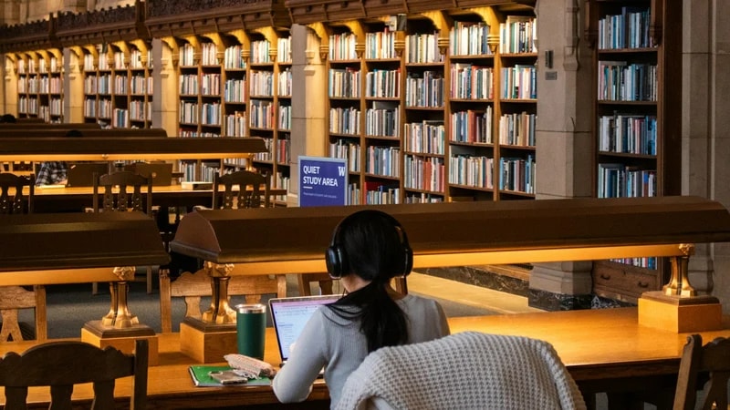 A student with headphones works on a laptop at a long wooden table in a quiet library study area
