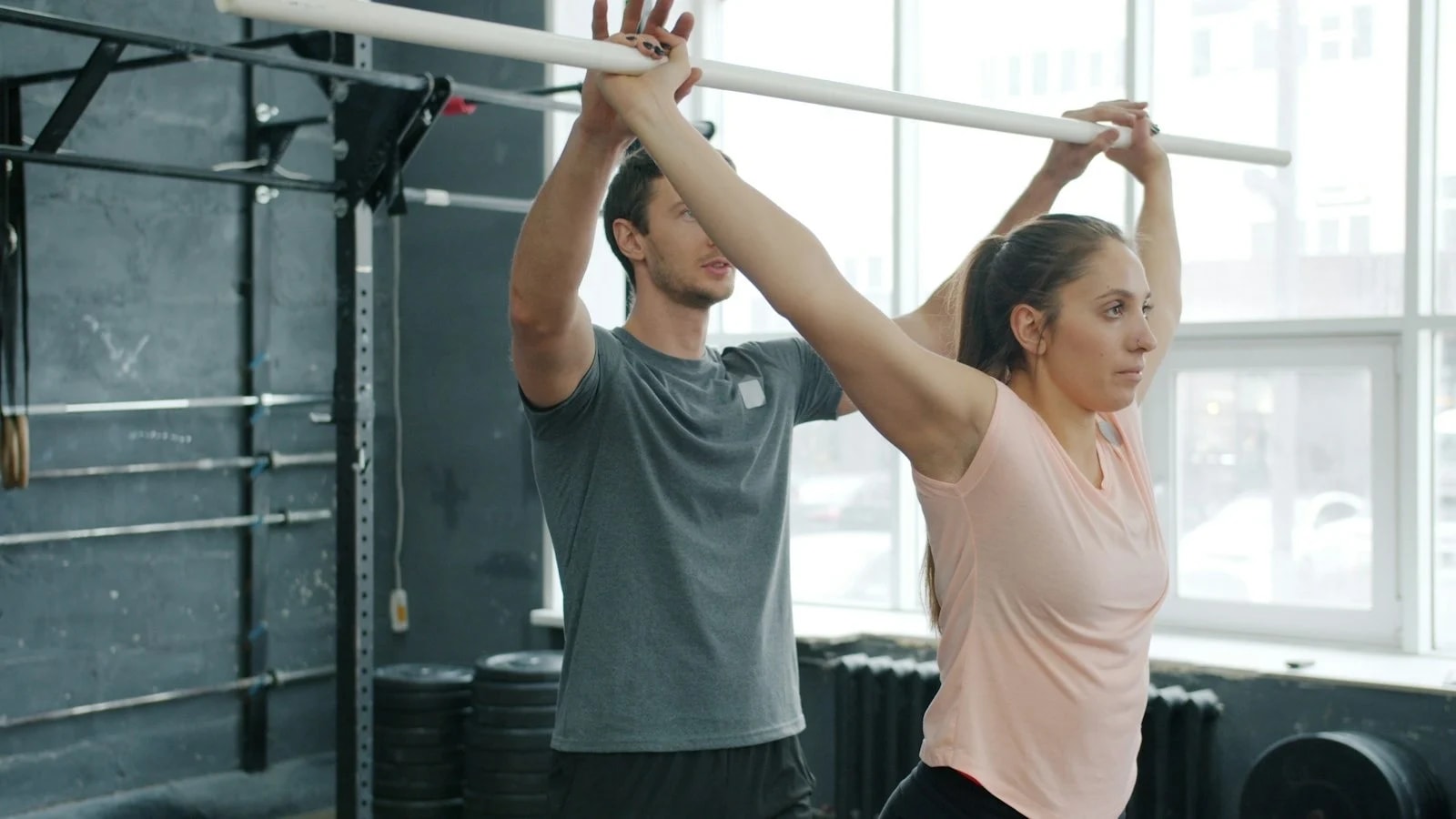 A coach standing behind a woman and guiding her as she presses a barbell overhead