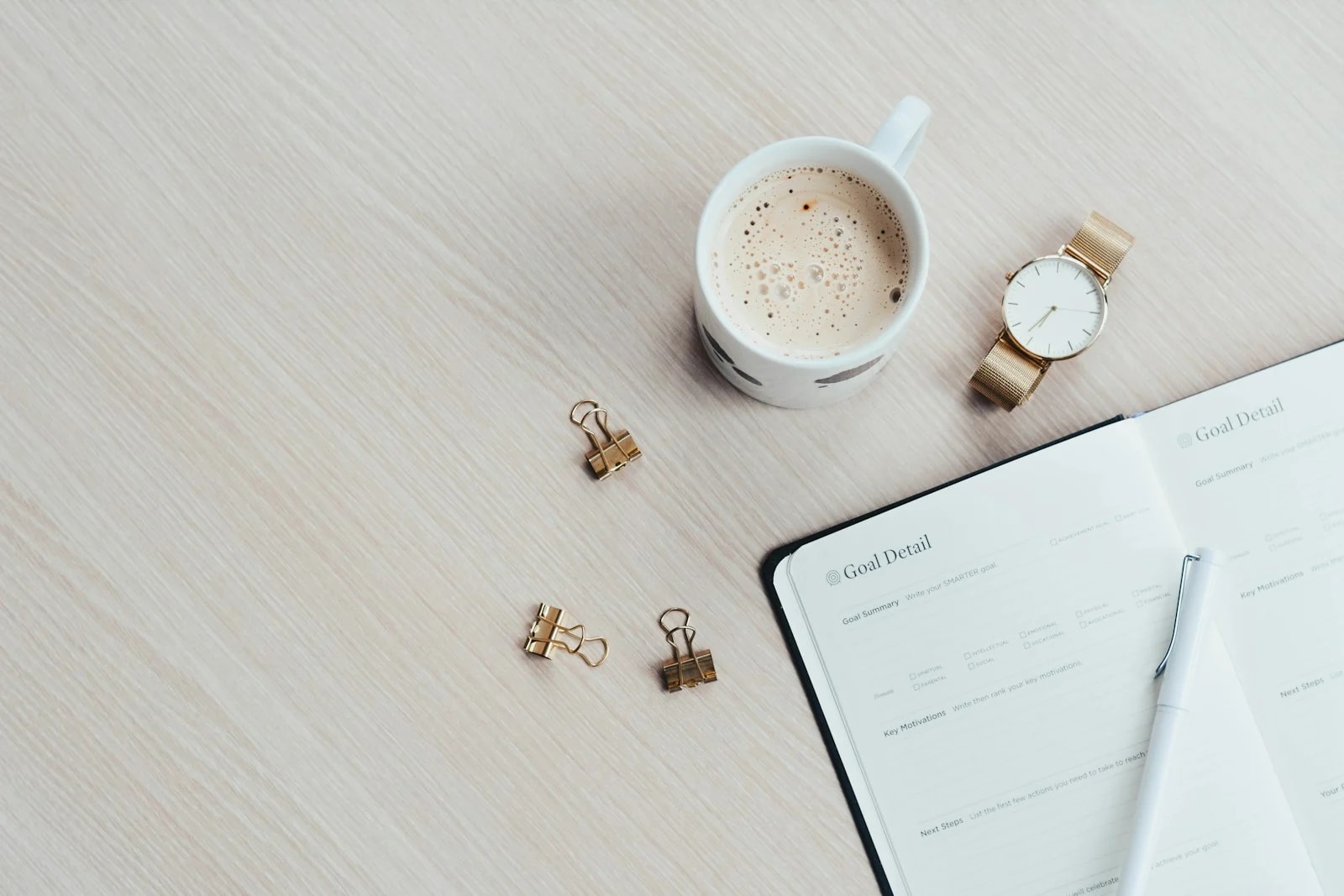 An open goal planner with a pen next to a cup of coffee and a gold watch on a light wooden desk