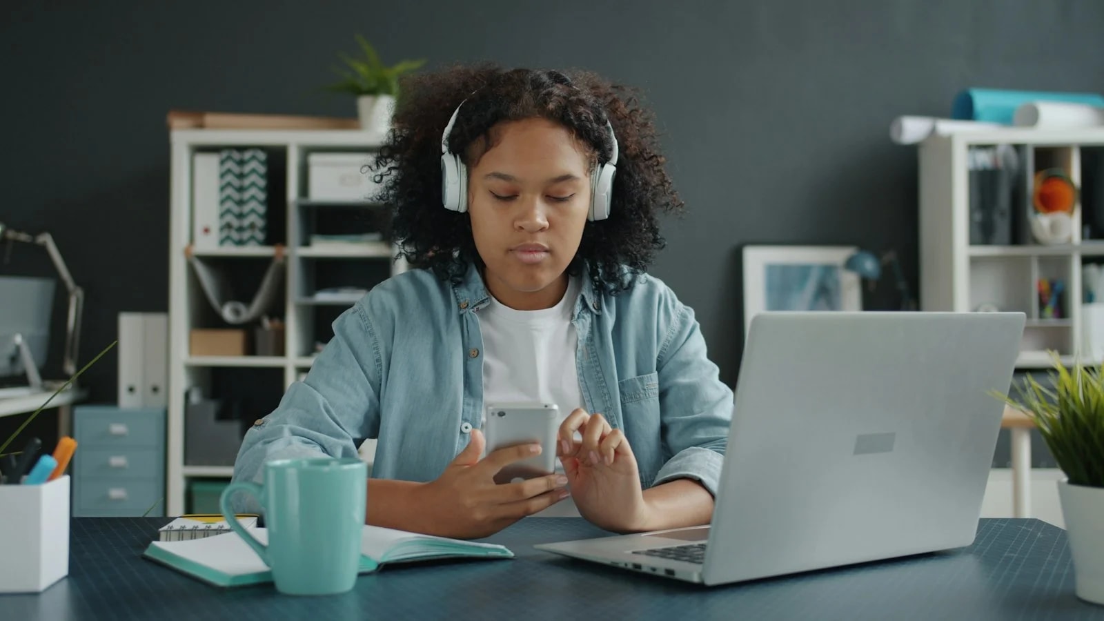 A young woman with headphones looks at her phone instead of the open laptop and notebook on her desk