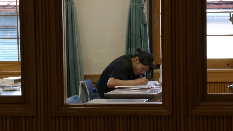 A student bent over a desk writing notes in a quiet library room, seen through a wooden window frame