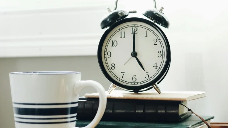 A black analog alarm clock on a stack of books next to a striped white mug, lit by a bright window