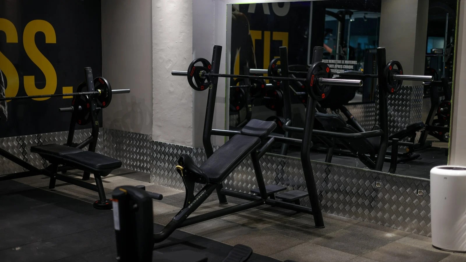 Una sala de musculación vacía con bancos, barras en soportes y un espejo en la pared