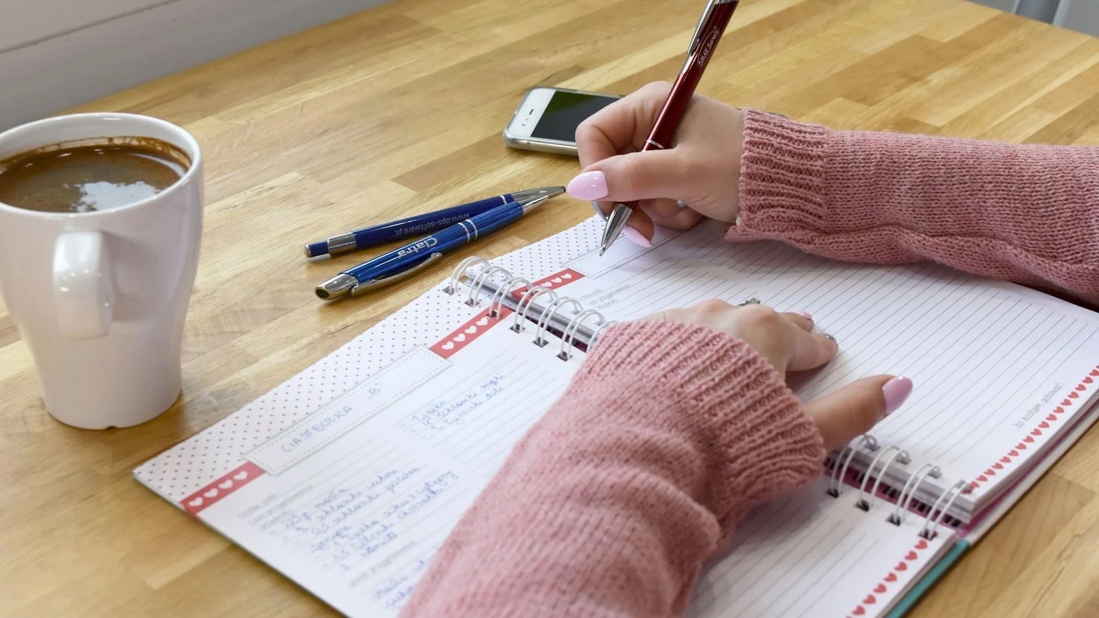 A person writing in a spiral planner at a wooden table next to a mug of coffee