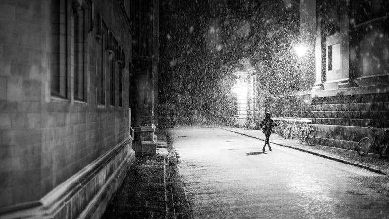 A person running down a narrow city street at night in heavy rain, in black and white