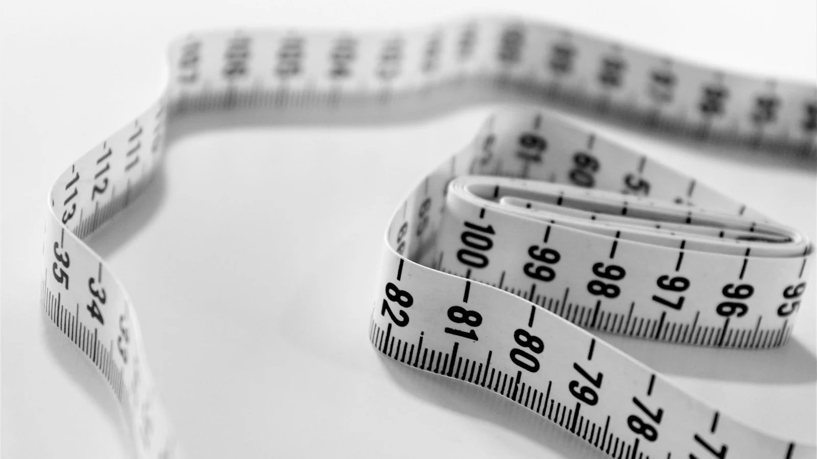 A white tailor's tape measure curled on a white surface, in black and white