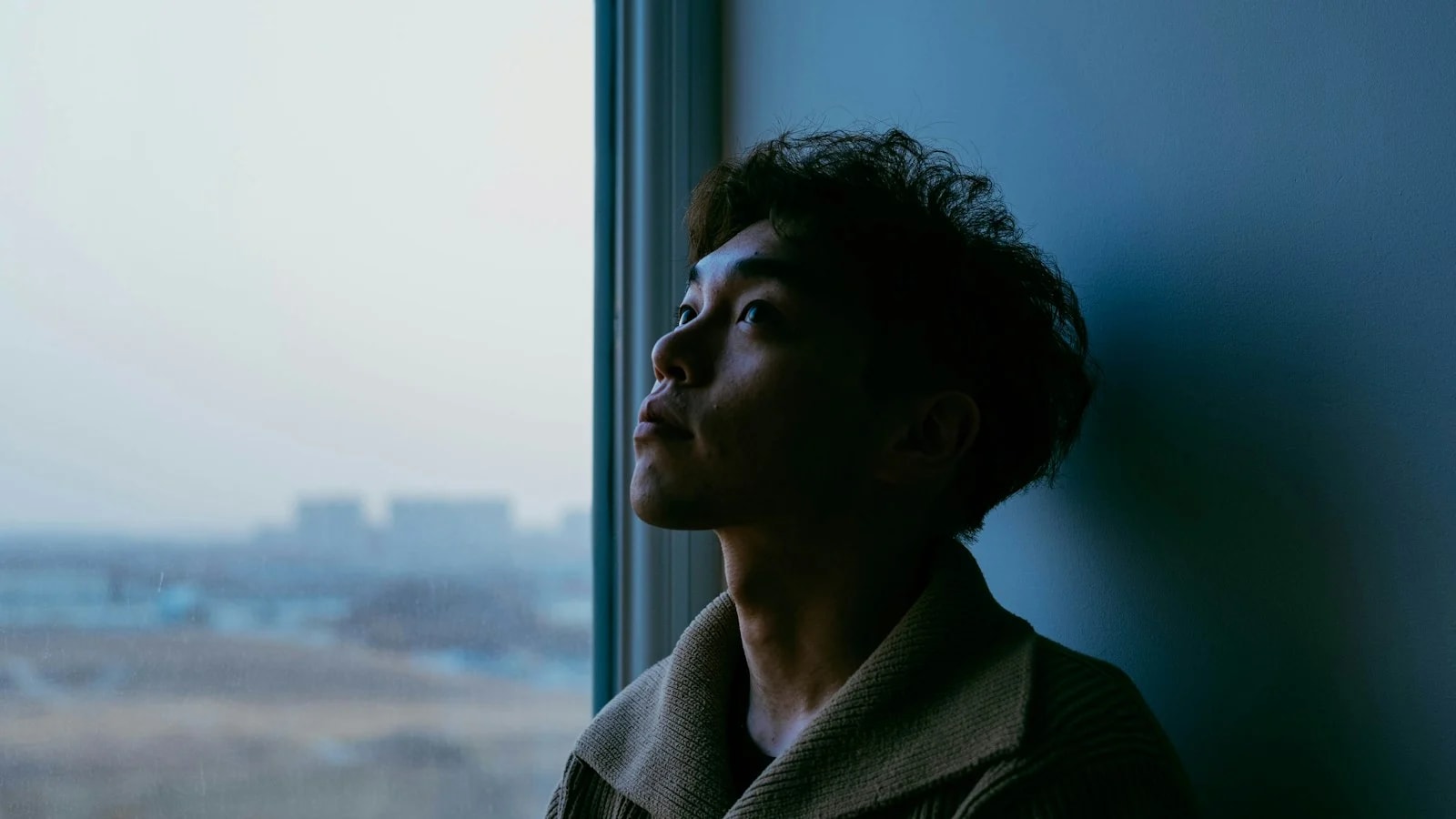 A young man leans against a wall next to a window and looks up, doing nothing, with a hazy city behind the glass