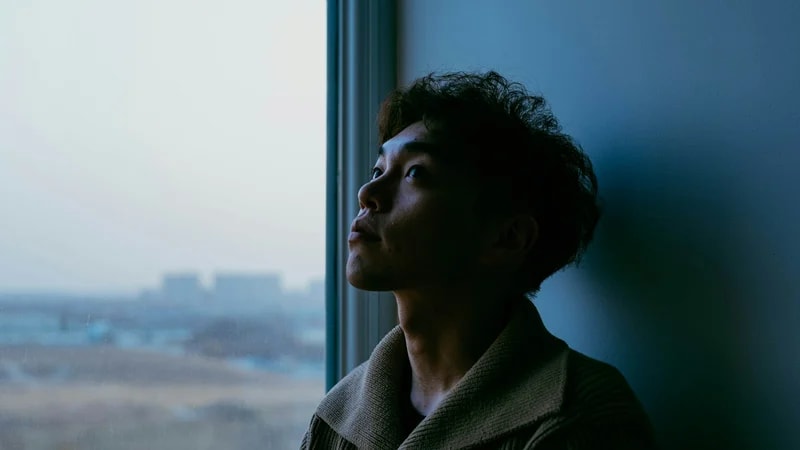 A young man leans against a wall next to a window and looks up, doing nothing, with a hazy city behind the glass
