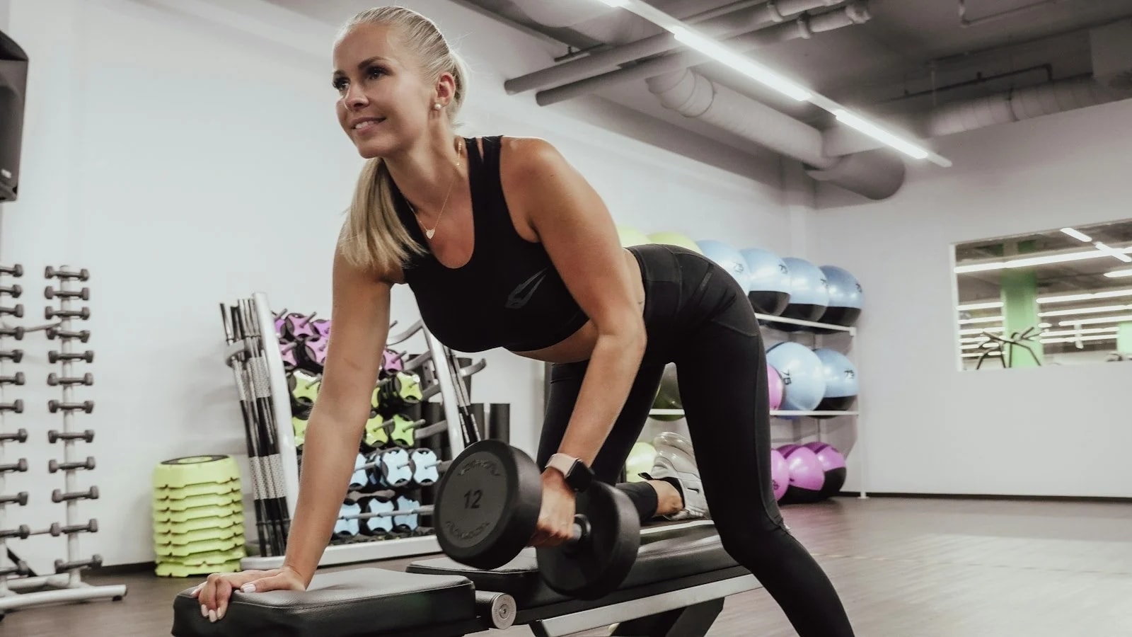 A woman doing a dumbbell row on a bench in a bright gym