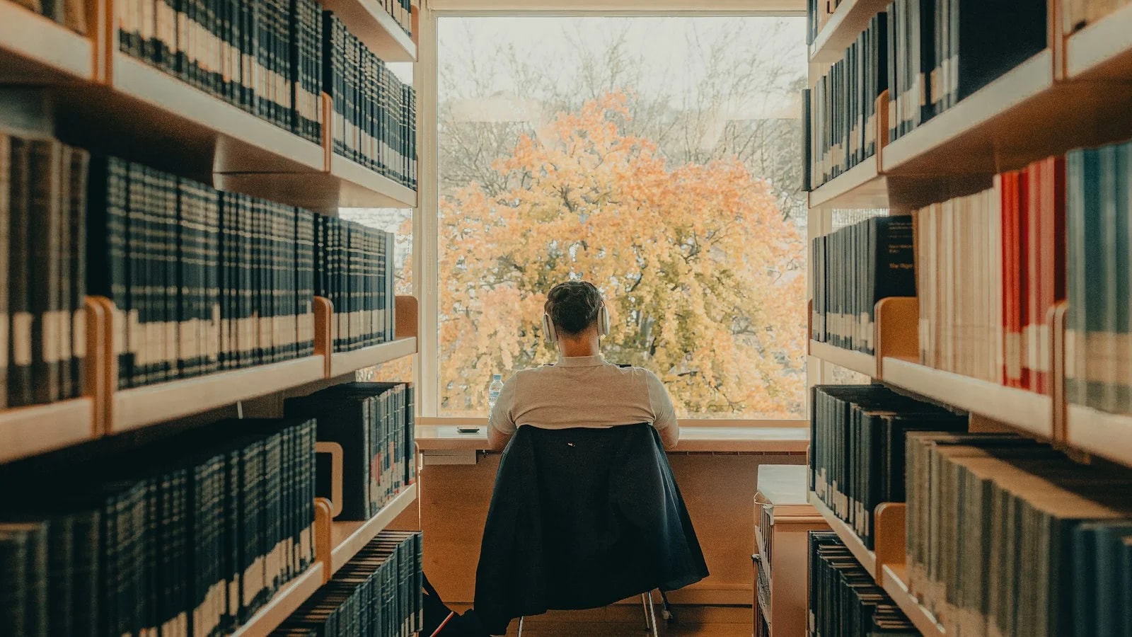 A student with headphones studies at a library desk facing a big window, between two rows of bookshelves, with an autumn tree outside