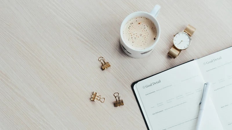 An open goal planner with a pen next to a cup of coffee and a gold watch on a light wooden desk