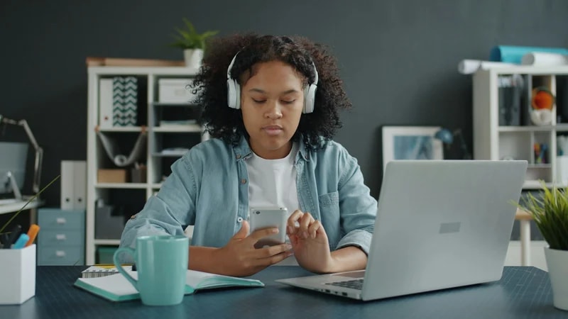 A young woman with headphones looks at her phone instead of the open laptop and notebook on her desk