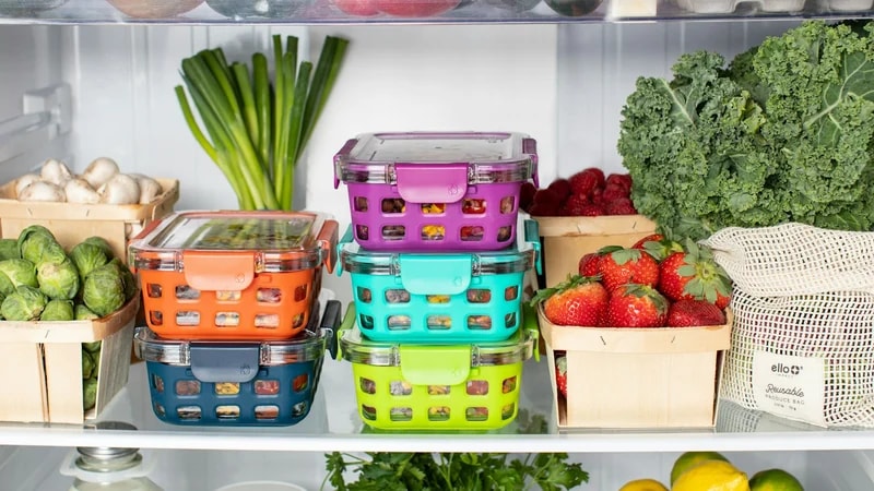 Stacked meal prep containers in a fridge next to strawberries, kale, mushrooms and Brussels sprouts