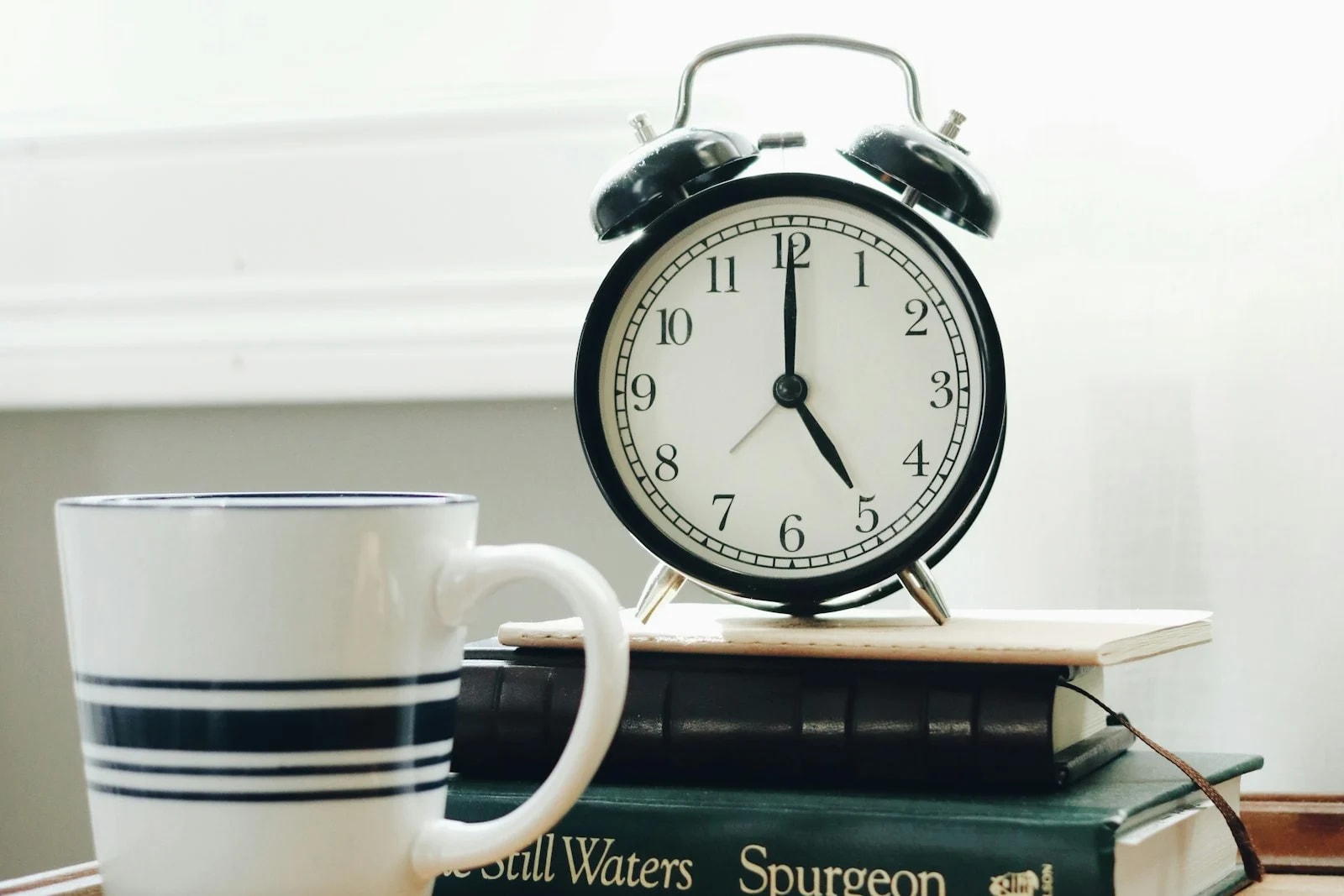 A black analog alarm clock on a stack of books next to a striped white mug, lit by a bright window